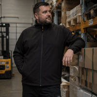 Mężczyzna w czarnej kurtce roboczej softshell JCB w magazynie z wózkiem widłowym w tle