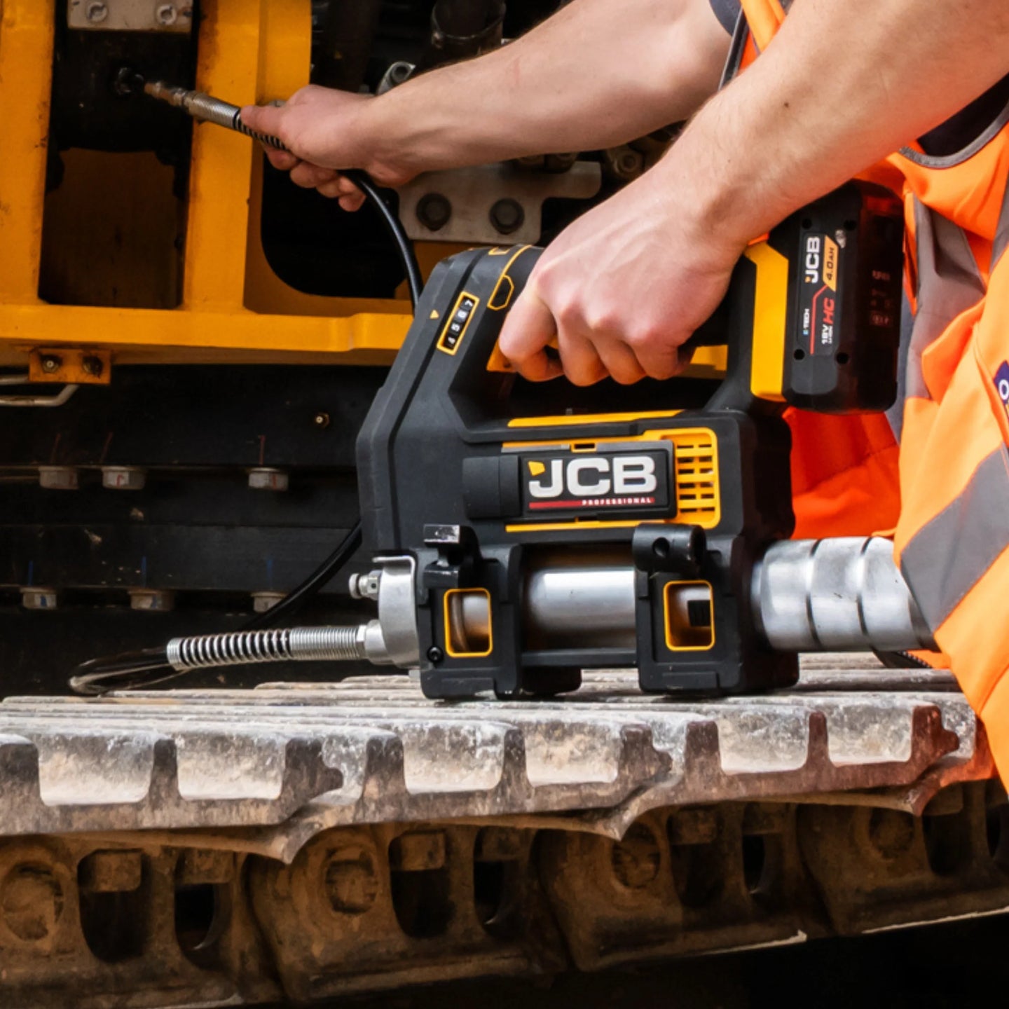 Smarownica akumulatorowa JCB 18V z zestawu JCB-18PKIT-5 trzymana w dłoniach nad gąsienicą maszyny napis Kontrola Dozowania