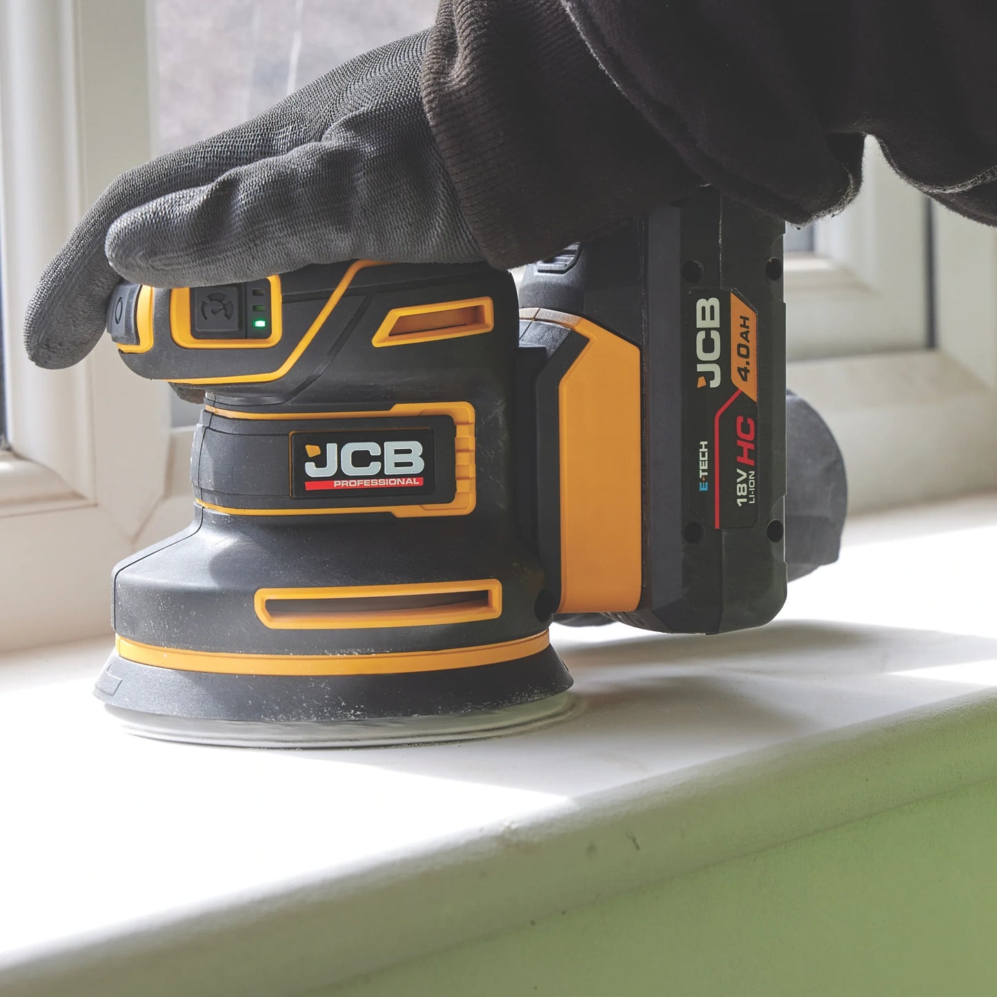 szlifierka mimośrodowa JCB Professional 18V podczas szlifowania drewna trzymana w rękawicy z tekstem Silnik bezszczotkowy